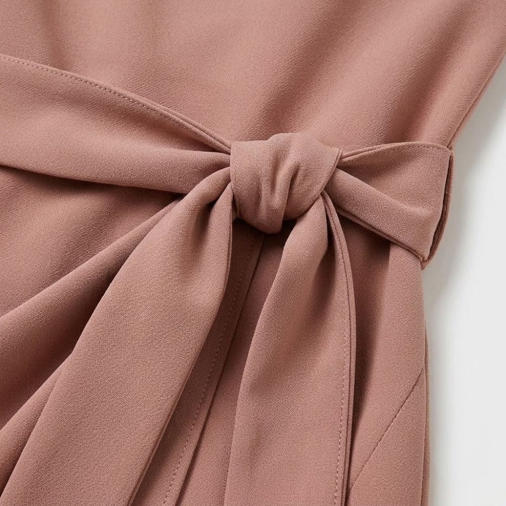 Detalhe de vestido curto em crepe rosa com laço ajustável na cintura e sobreposição frontal elegante.