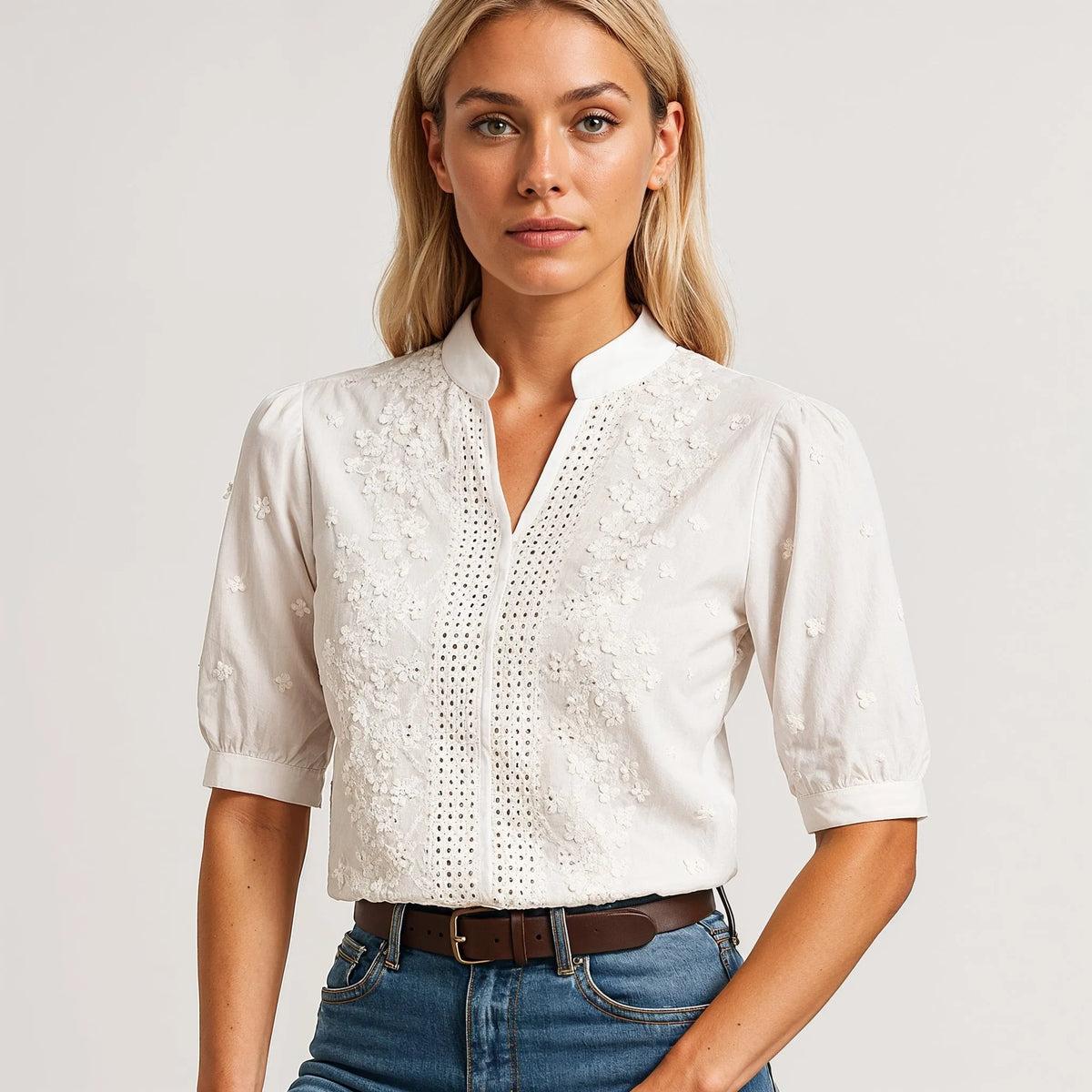 Blusa branca de algodão com bordado floral, detalhes perfurados, gola curta e decote em V, usada com calças de ganga.